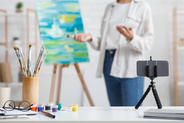 Zeitgenössische Kunst, Hobbys und Online-Unterricht zu Hause. Smartphone, Farbe und Pinsel im Fokus, afrikanisch-amerikanische Künstlerin malt im Wohnzimmer - Foto, Bild