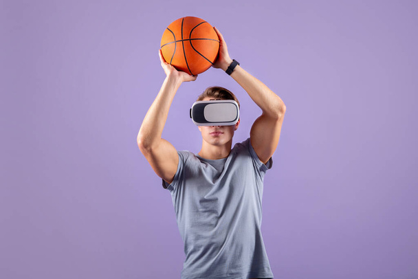 Концепция виртуального спорта. Молодой мускулистый парень, играющий в баскетбол в VR гарнитуре на фоне фиолетовой студии - Фото, изображение