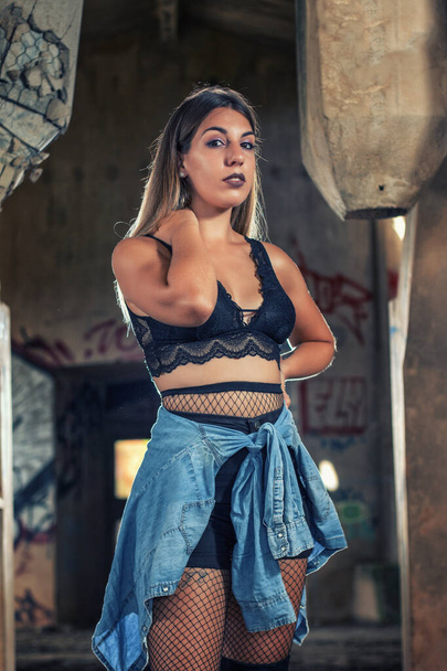 Young girl posing in a fashion way on an abandoned factory building using a blue denim shirt, black top and short jeans. - Fotografie, Obrázek
