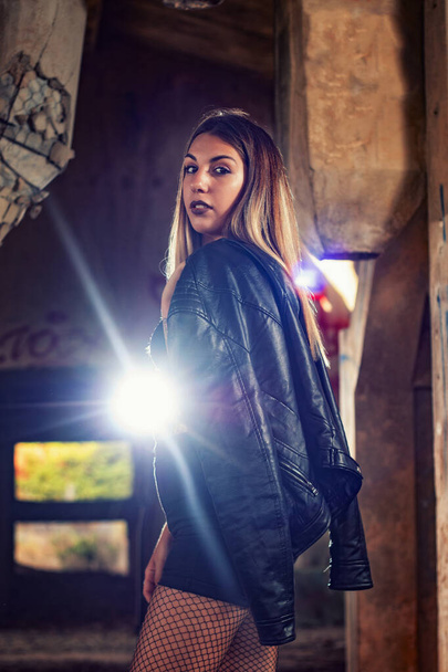 Menina posando de uma forma de moda em um prédio de fábrica abandonada usando uma jaqueta de couro, top preto e jeans curtos. - Foto, Imagem