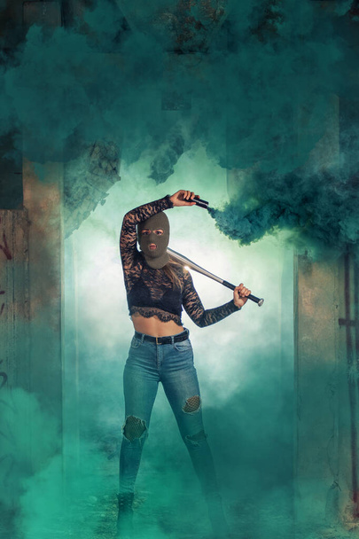 Chica peligrosa con murciélago y pasamontañas en una fábrica abandonada usando una granada de humo. - Foto, Imagen