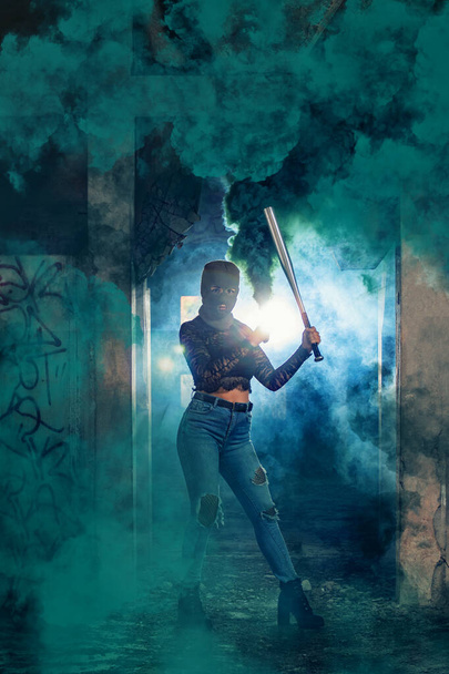 Небезпечна дівчина з кажаном і балаклавою на покинутому заводі, використовуючи димову гранату
. - Фото, зображення