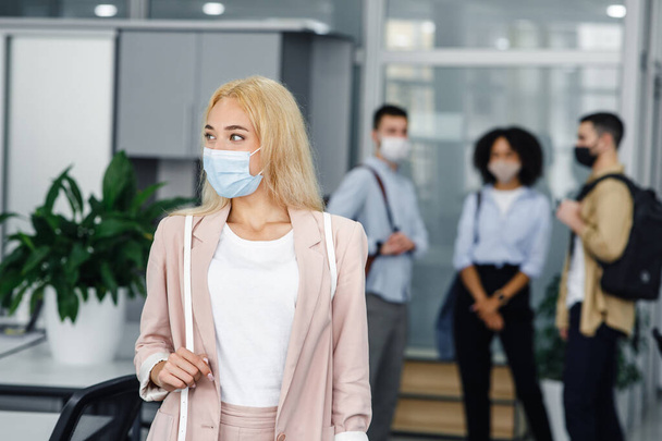 Workers in modern office hallway return to work after quarantine. Focus on beautiful blonde woman in business suit and protective mask - Zdjęcie, obraz