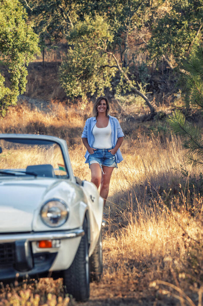 田舎を歩く白いコンバーチブルヴィンテージ車を持つ女性. - 写真・画像