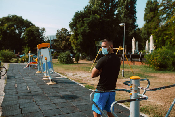 Vista trasera de un joven atleta masculino muscular con una mascarilla de pie junto a un dispositivo en un gimnasio al aire libre. Hombre en formación. Protección contra el Coronavirus COVID - 19 - Foto, imagen