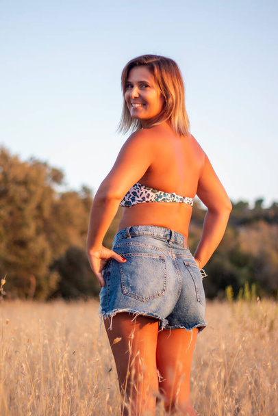 Mooi meisje met bikinitop en jeans op het platteland met droge gras vegetatie. - Foto, afbeelding