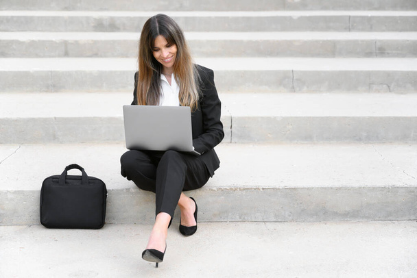 Beautiful businesswoman in suit, using a laptop sitting on stairs outdoors - Photo, Image