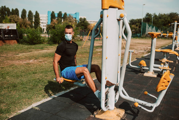 Fiatal kaukázusi atléta, arcmaszkos vonattal a karján és a lábán egy szabadtéri edzőteremben. Izmos sportoló a szabadban. COVID - 19 koronavírus elleni védelem - Fotó, kép