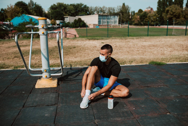 Fitter junger männlicher kaukasischer Athlet mit Gesichtsmaske sitzt und bindet Schnürsenkel in einer Turnhalle in der Natur. Muskelprotz im Freien. COVID - 19 Coronavirus-Schutz - Foto, Bild