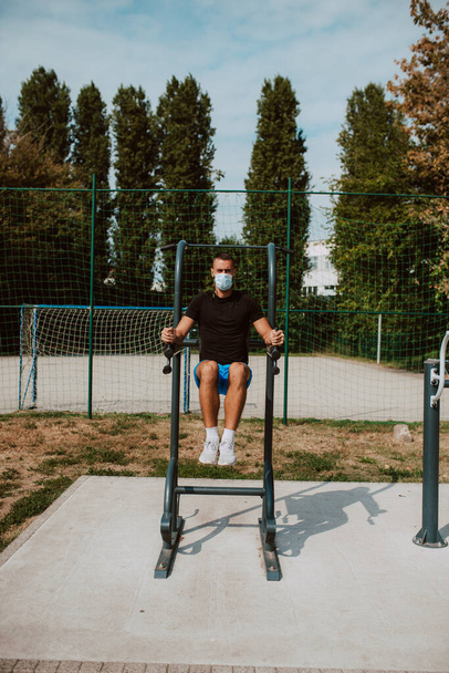 Fit jonge mannelijke blanke atleet met gezichtsmasker treinen op arm en been apparatuur in een outdoor fitnessruimte. Spiersporter buiten. COVID - 19 bescherming tegen coronavirus - Foto, afbeelding