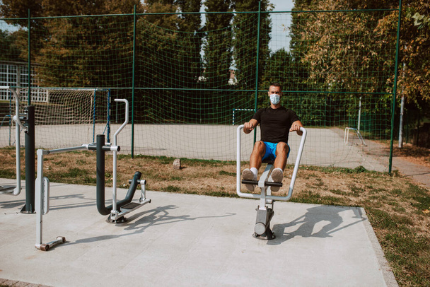 Adatto a giovani atleti caucasici maschi con maschera facciale si allena su attrezzature per braccia e gambe in una palestra all'aperto. Atleta muscolare all'aperto. COVID - 19 protezione coronavirus - Foto, immagini
