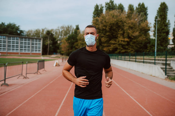 A shot of a young male caucasian athlete  with face mask training on a racetrack. The sprinter runs on athletic all-weather running track. COVID - 19 coronavirus protection - Φωτογραφία, εικόνα