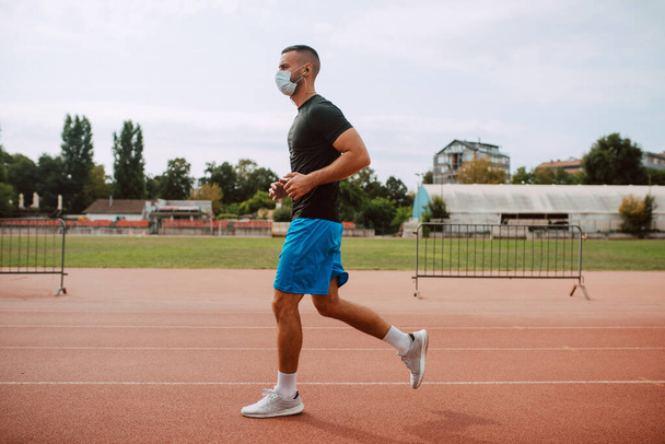 Een foto van een jonge blanke atleet met gezichtsmasker training op een racebaan. De sprinter loopt op een atletisch hardloopparcours. COVID - 19 bescherming tegen coronavirus - Foto, afbeelding