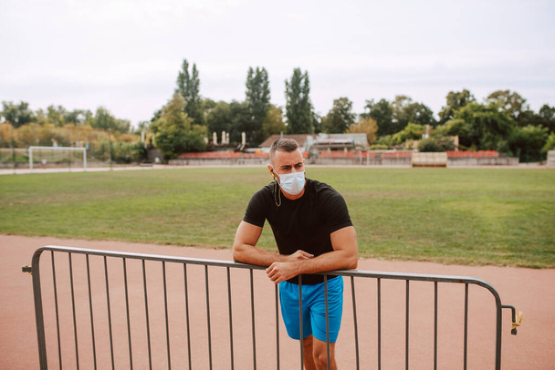 Futbol sahasının yanındaki çitlere yaslanmış, yüzü maskeli, beyaz, genç bir erkek sporcu. COVID - 19 koronavirüs koruması - Fotoğraf, Görsel