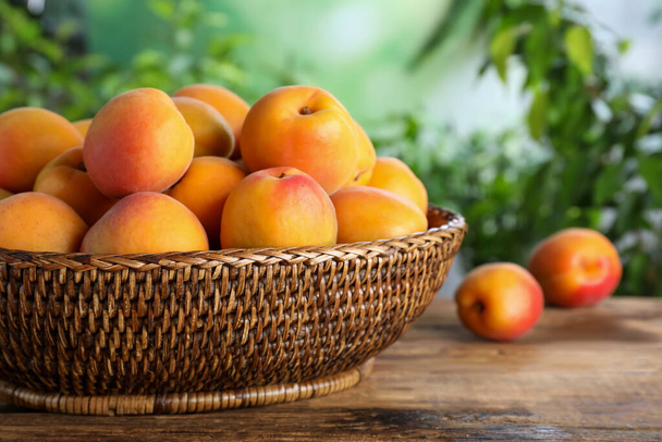 Many fresh ripe apricots in wicker bowl on wooden table against blurred background - Foto, immagini