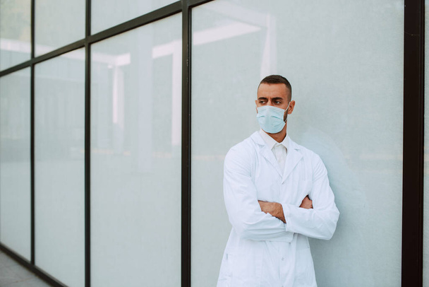 Ritratto di un giovane medico caucasico in uniforme bianca con una maschera protettiva sul volto di fronte all'edificio. Protezione contro il COVID - 19 coronavirus - Foto, immagini