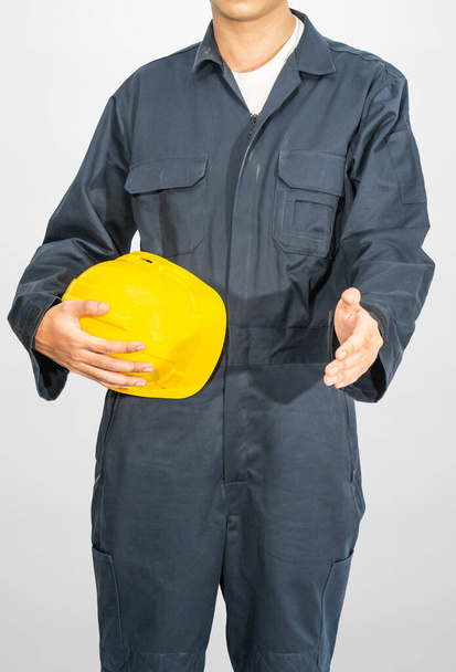 灰色の背景に隔離された黄色の硬い帽子を保持する青いカバーオールに立つ労働者 - 写真・画像