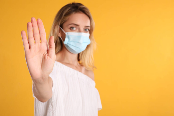 Woman in protective face mask showing stop gesture on yellow background, focus on hand. Prevent spreading of coronavirus - Foto, Bild