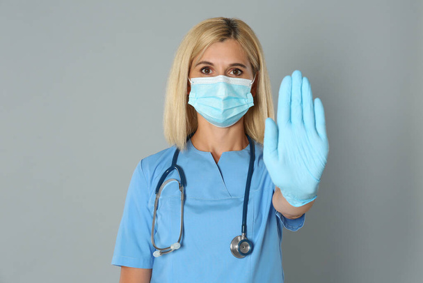 Doctor in protective mask showing stop gesture on grey background. Prevent spreading of coronavirus - Φωτογραφία, εικόνα