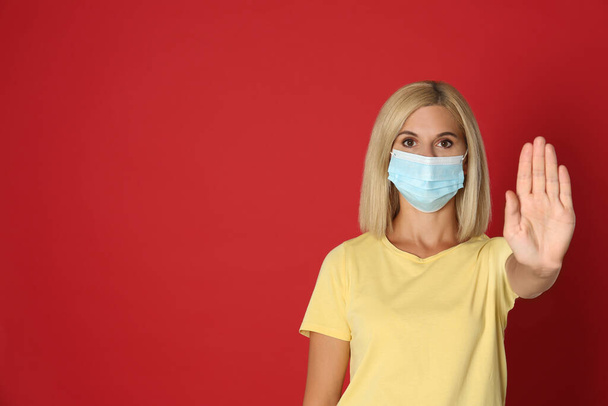 Woman in protective mask showing stop gesture on red background, space for text. Prevent spreading of coronavirus - Fotografie, Obrázek