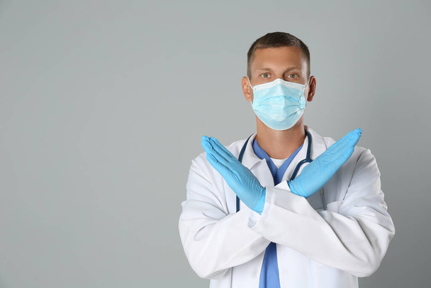 Doctor in protective mask showing stop gesture on grey background, space for text. Prevent spreading of coronavirus - Фото, зображення