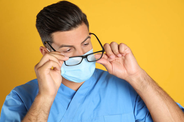 Doctor wiping foggy glasses caused by wearing medical mask on yellow background - Foto, Imagem