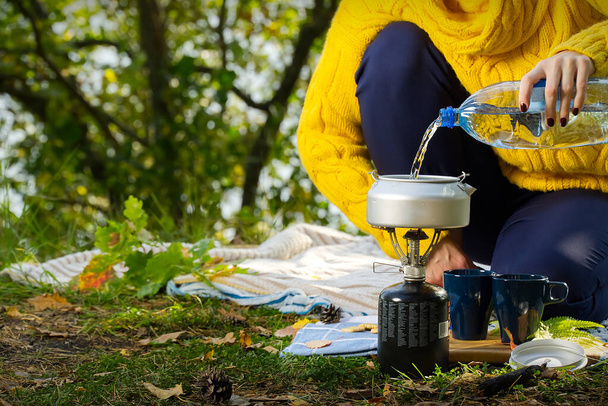 jong mooi meisje in een gele trui maakt koffie in het bos op een gasbrander. Koffie zetten op een primus fornuis in het herfstbos, stap voor stap. Kamperen in herfstbos concept - Foto, afbeelding