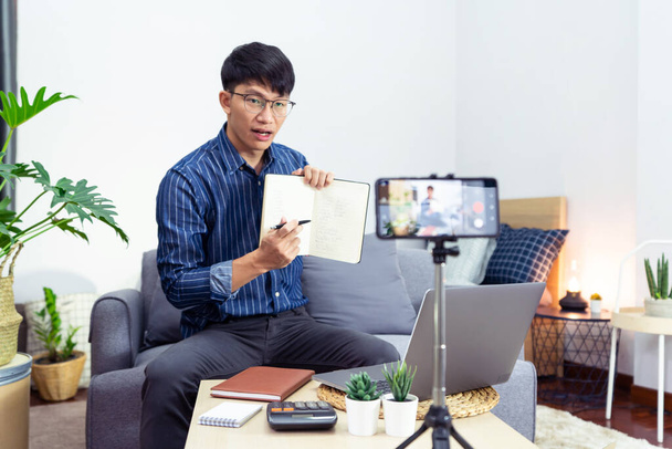ノートブックでノートを書くアジア人男性ホームレクチャー研究でラップトップを介してウェビナービデオコースの研究を見てオンラインで、 E-Learningの概念 - 写真・画像