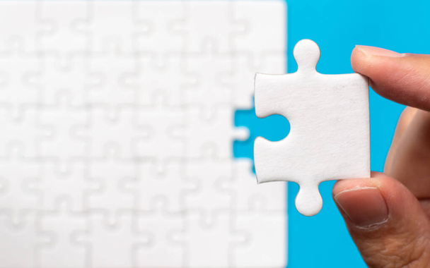 Hand holding piece of white jigsaw puzzle blurred background. Team business success partnership or teamwork. - Photo, Image