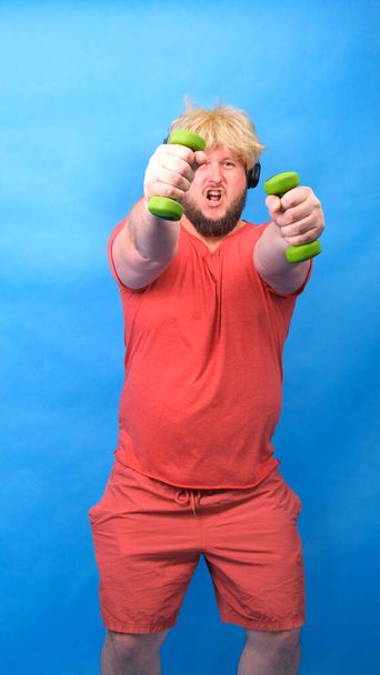 Funny chubby freak man in headphones and a wig in a pink t-shirt is engaged in green dumbbells and laughs on a blue background, vertical - Фото, изображение