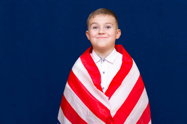 Boldog fiatal kaukázusi amerikai fiú gyerek iskolás fiú becsomagolja magát egy amerikai zászló és belenéz a kamera kék háttér. - Fotó, kép