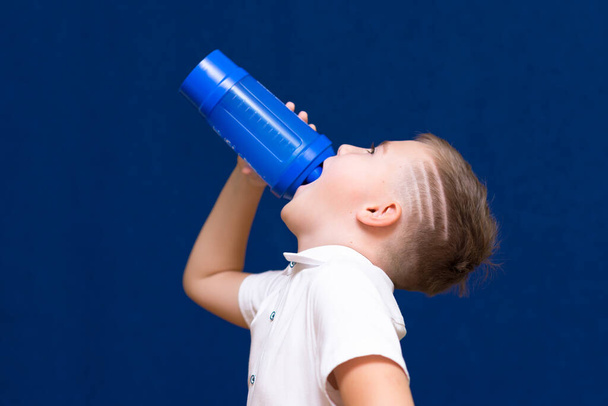 Side view close up Young caucasian boy wearing white t-shirt holding water bottle kid drinking on blue - Photo, image
