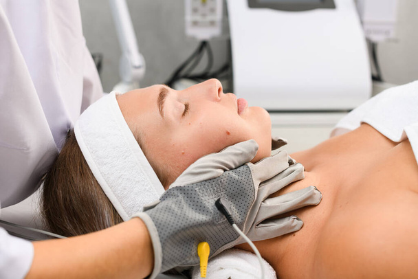 Sprzęt profesjonalny kosmetolog dotknąć twarz klienta z szarymi mikroprądowymi rękawiczkami do myostymulacji skóry terapii zbliżenie, widok z boku - Zdjęcie, obraz