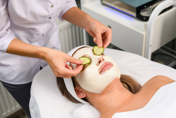 Kosmetikerin trägt natürliche weiße feuchtigkeitsspendende Gesichtsmaske auf und erfrischt Augenpartie mit Gurke, Pflegekonzept, professionelle Behandlung für liegende und entspannende Patientin - Foto, Bild