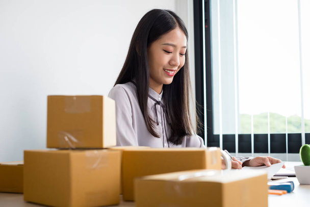 Jonge Aziatische vrouw ondernemer met veel pakket dozen op de tafel gelukkig online verkoop baan, gebruik maken van uw laptop, krijgen een bestelling van klanten, notities te maken, en regelingen voor de levering per post. - Foto, afbeelding