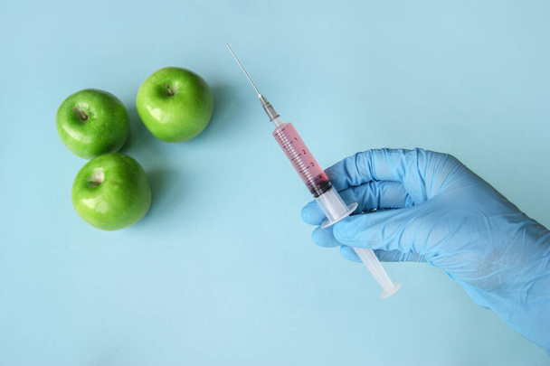 Green apple and syringe with GMO in hands on a blue background. GMO concept with vegetables and fruits. - Photo, Image