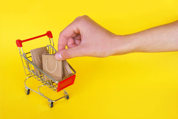 Mini carro y mano femenina con pequeñas bolsas artesanales sobre fondo amarillo. El concepto de compras en las tiendas y en Internet. - Foto, Imagen