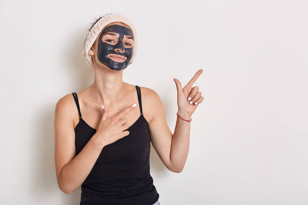 Portrait of beautiful girl with hairband on her head, has mud facial mask, looks satisfied at camera, pointing with forefingers aside, standing against white background. - Фото, зображення