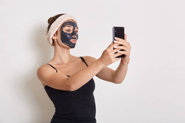 Portrait of girl with black cosmetic mask on her face holding modern smart phone in hand and looking at device screen, making selfie isolated over white background. - Foto, Imagen
