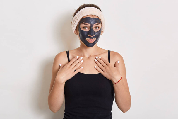 Adorable female with hairband looking at camera, posing in sleeveless t shirt, doing beauty procedures at come, applies mud black mask on face for moisturising. - 写真・画像