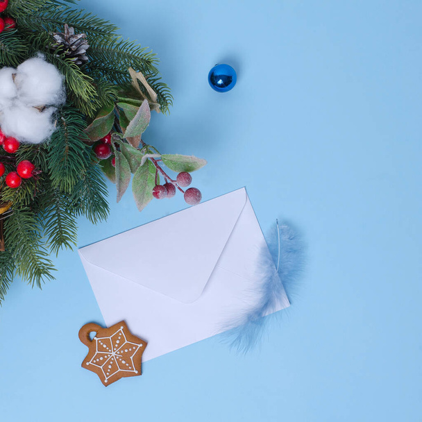 New Year's concept with a Christmas tree and Christmas decorations, a white envelope for a letter on a blue background lies nearby. there is a gingerbread and a feather on the envelope. Flat lay, copy space - Фото, изображение