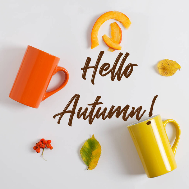 Witaj, Autumn! napis na białym tle pomiędzy pomarańczowymi i żółtymi filiżankami herbaty. Jesienne prezenty, liście drzew, popiół górski i kawałki dyni. Skupienie selektywne, płaski układ - Zdjęcie, obraz