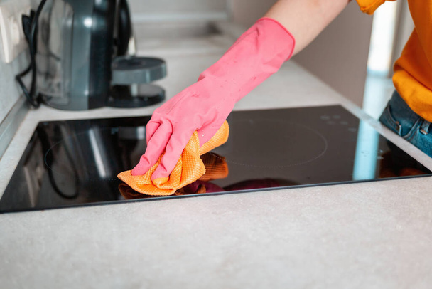 Μια γυναίκα με λαστιχένια γάντια σκουπίζει μια επαγωγική κουζίνα με ένα πανί. Χέρι κοντά. - Φωτογραφία, εικόνα
