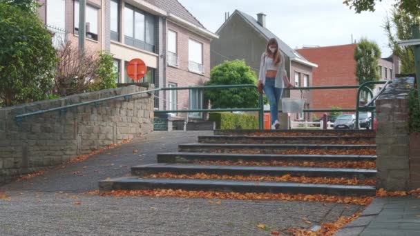 μια νεαρή γυναίκα σε περιστασιακό φόρεμα κρατώντας ένα καλάθι με τα πόδια κάτω από τις σκάλες σε δημόσιο δρόμο - Πλάνα, βίντεο