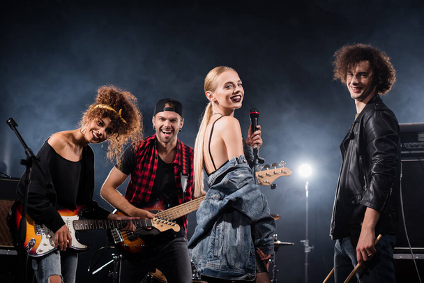 KYIV, UKRAINE - AUGUST 25, 2020: Happy rock band vocalist standing near curly drummer and smiling guitarists with backlit on background - Photo, image