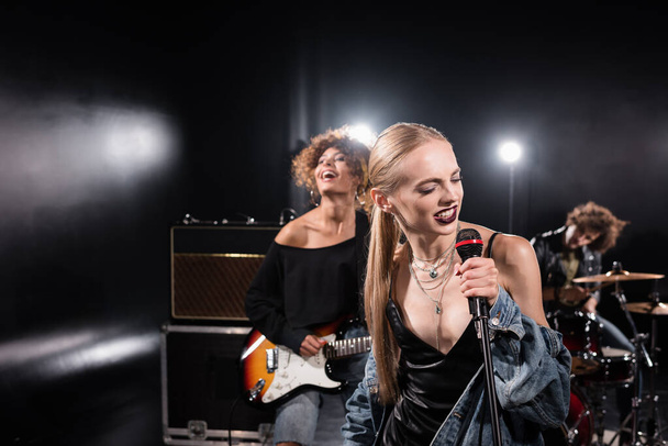 KIEW, UKRAINE - 25. AUGUST 2020: Blonde Frau singt mit Mikrofon neben Gitarrist mit hinterleuchtetem und verschwommenem Schlagzeuger im Hintergrund - Foto, Bild