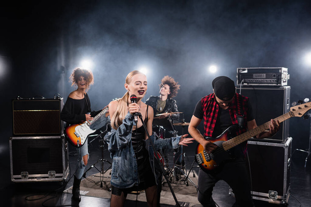 KYIV, UKRAINE - AUGUST 25, 2020: Woman with closed eyes singing while holding microphone rack near rock band musicians on blurred background - Foto, afbeelding