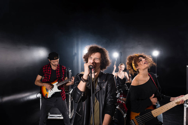 KYIV, UKRAINE - AUGUST 25, 2020: Curly vocalist with closed eyes singing in microphone near guitarists with backlit and female drummer on background - Photo, Image