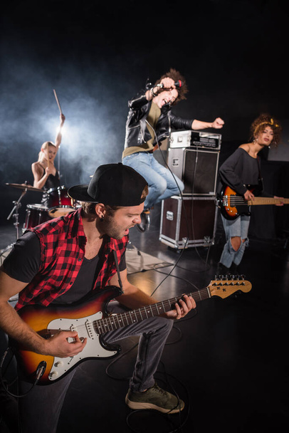 KYIV, UCRANIA - 25 de agosto de 2020: Músico de la banda de rock tocando la guitarra eléctrica, mientras que el vocalista rizado salta y grita en el micrófono sobre un fondo borroso - Foto, Imagen