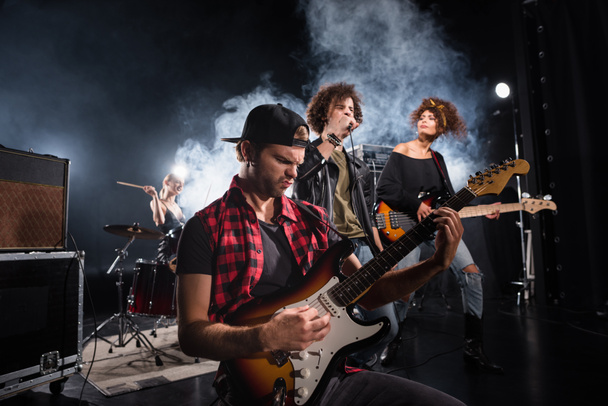 KIEW, UKRAINE - 25. AUGUST 2020: Rockband-Musiker mit Bassgitarre sitzt neben lockigem Sänger und Gitarristen mit Rauch und Schlagzeugerin im Hintergrund - Foto, Bild